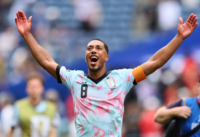 Soccer player celebrates with arms raised, wearing a light blue and pink jersey with number 8 and an orange captain's armband.