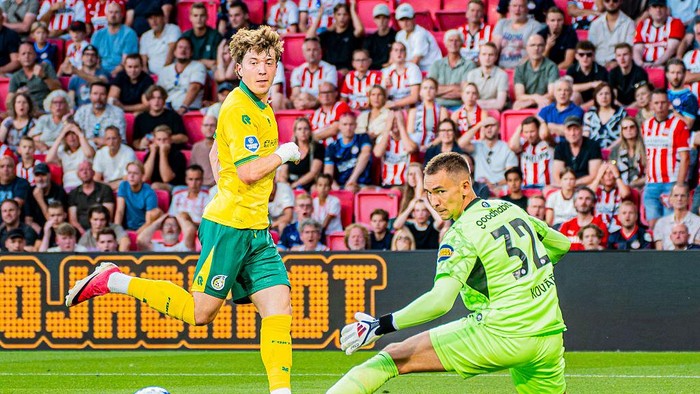 Forward in a yellow kit kicks the ball as the neon-green goalkeeper dives to save, with a packed stands of fans behind.