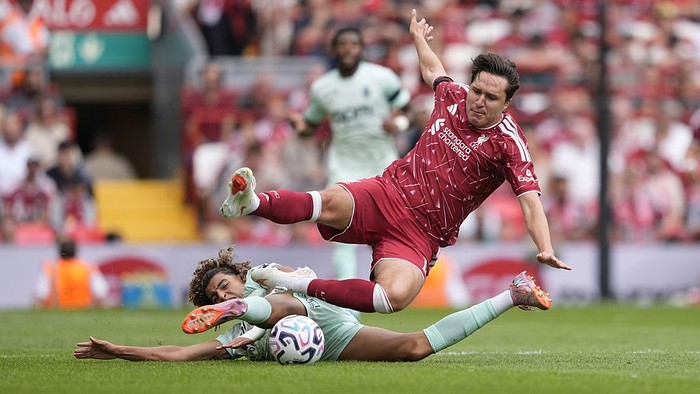 Red-jersey soccer player leaps over a sliding opponent in light blue as they contest the ball on a green pitch.