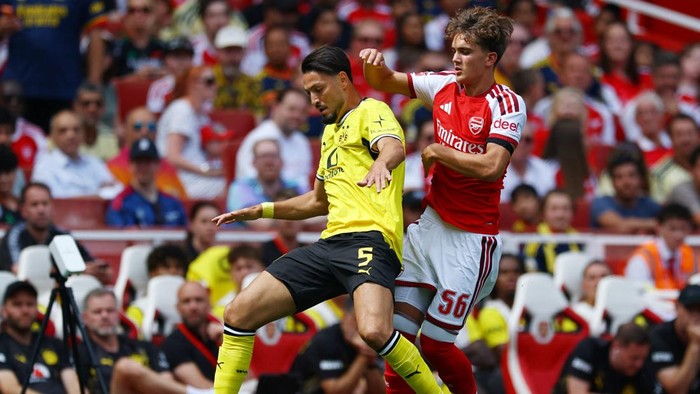 Two soccer players in a duel for the ball during a match, one in yellow/black and the other in red/white, with fans in the stands behind them.