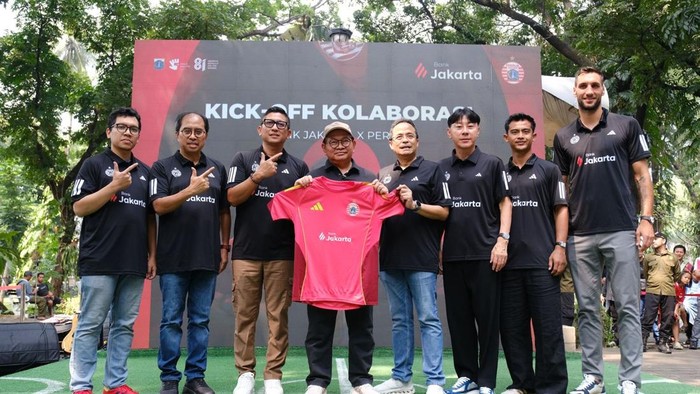 Eight men in matching black jerseys pose on a stage outdoors, holding a pink soccer jersey during a 'Kickoff Kolaborasi' event.