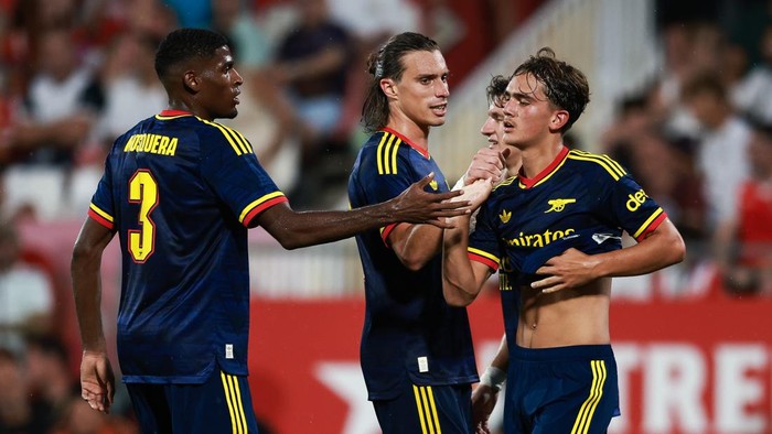 Three footballers in blue kits celebrate a goal on the pitch, one grabbing another's jersey in a group hug moment on the field.