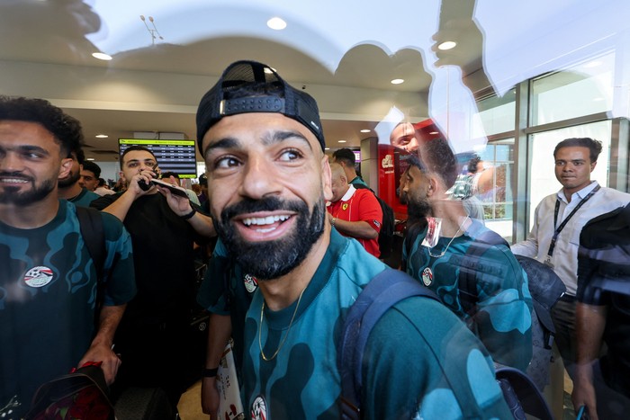 Smiling man with a beard in a teal jersey and backward cap among teammates at an airport arrivals area.