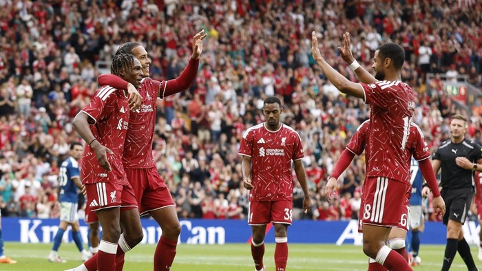 Liverpool FC players celebrate a goal on the pitch as fans cheer in the stands behind them.