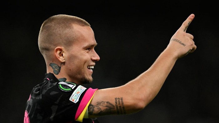 Male footballer with a shaved head smiling and pointing upward with his right hand, showing colorful tattoos on his arm and neck tattoo in a black jersey.
