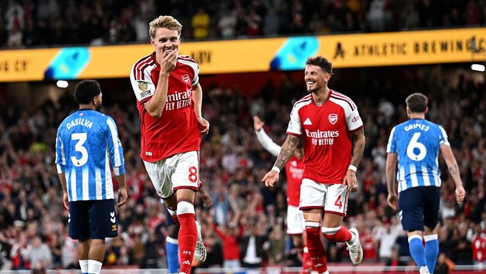 Arsenal players celebrate a goal on the pitch, wearing red jerseys and white shorts, with blue-and-white striped opponents nearby.