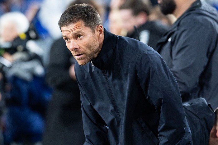 Man in a dark jacket bends forward on the sideline during a sports game, watching intently in a crowded stadium.