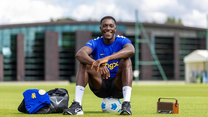 Young male soccer player in a blue jersey sits on the grass with a ball under his feet, smiling, flanked by a sports bag and a portable radio.