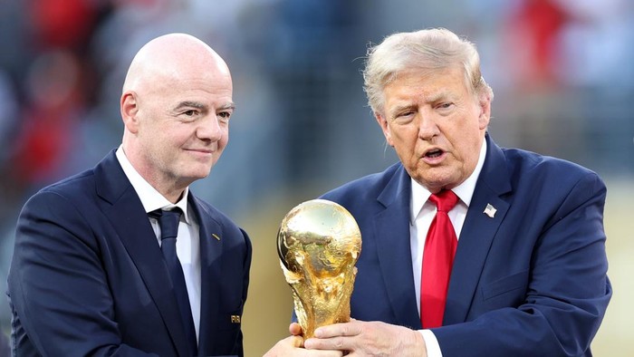 Two men in dark suits hold a shiny gold trophy outdoors, one wearing a red tie, faces toward the camera.