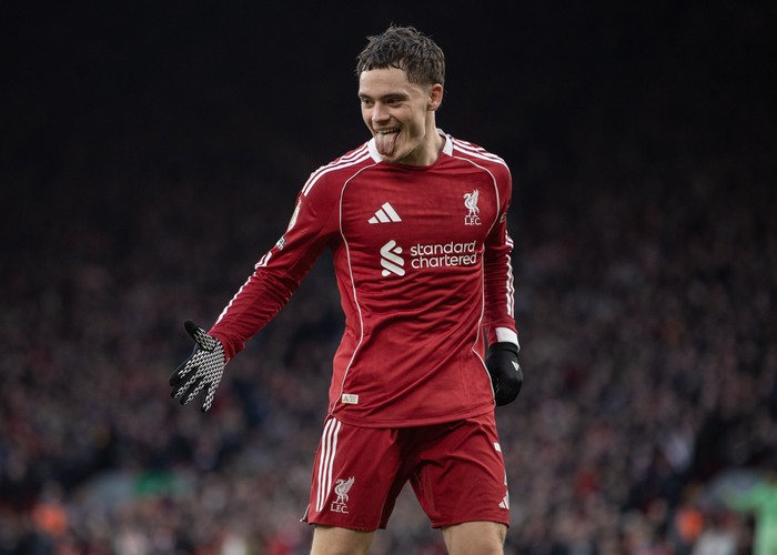 Soccer player in a red Liverpool jersey celebrates on the field with a smile and tongue out, wearing gloves.