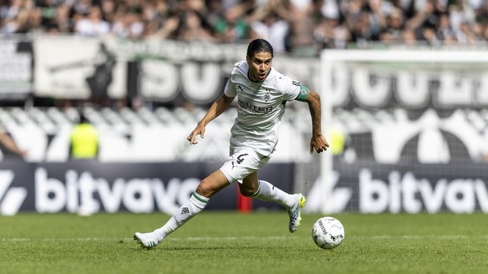 Soccer player in a white uniform dribbles the ball on a green pitch during a match, with spectators in the stands in the background.