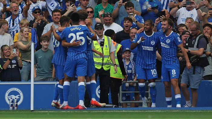 Blue soccer players celebrate a goal near the sideline as excited fans in the stands cheer and film the moment.