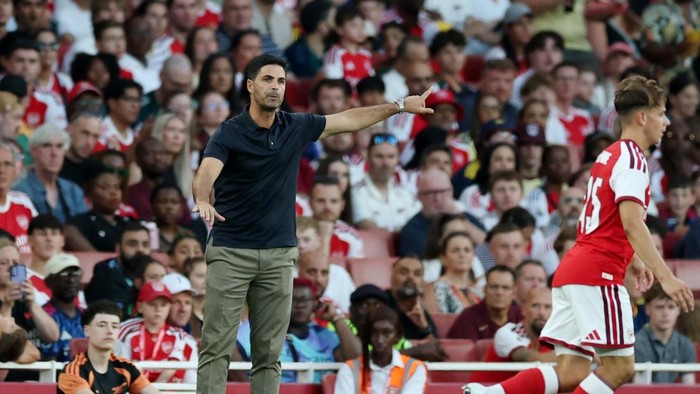 Soccer coach on the sideline gesturing with his right arm as a player in red and white runs by, stadium stands full of spectators in the background.
