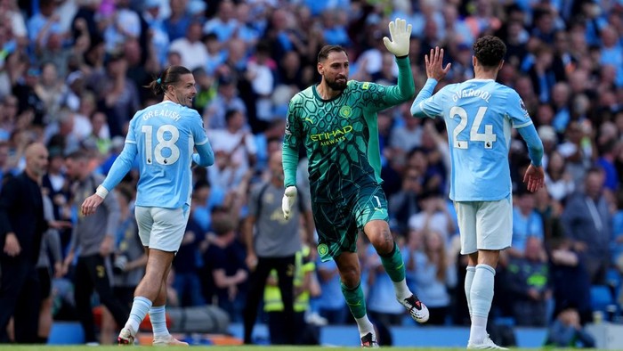 Manchester City players celebrate a goal on the pitch, with the goalkeeper in green raising a hand between two teammates wearing numbers 18 and 24.
