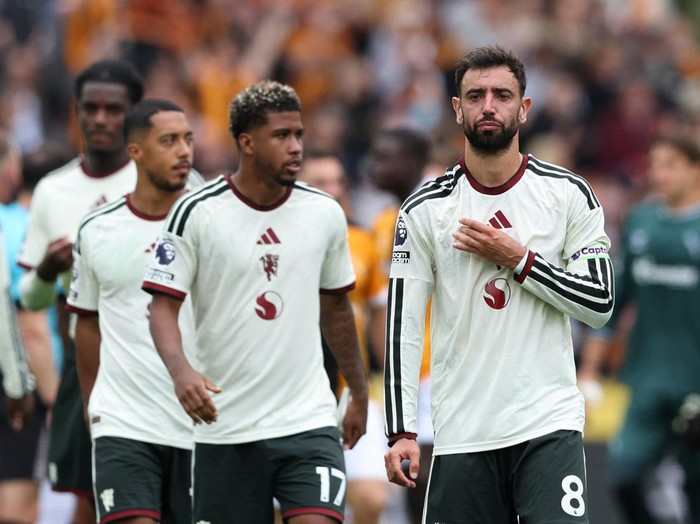 Three football players in white jerseys and dark shorts walk off the pitch together, with a blurred crowd in the background.