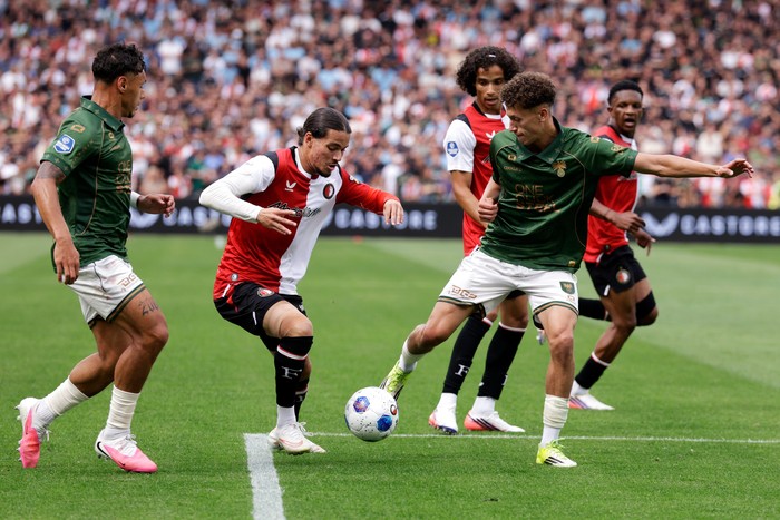 Soccer players from white-red and green teams contest the ball on a green pitch with a packed crowd behind.