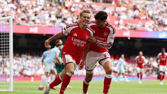 Arsenal teammates celebrate a goal on the pitch, wearing red jerseys with Emirates Fly Better, in front of cheering crowds.