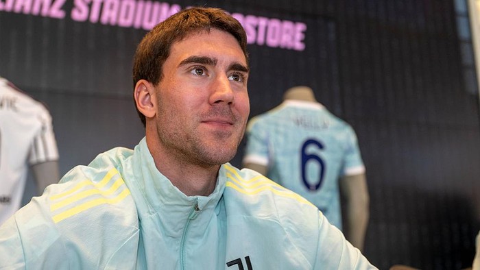 Portrait of a man in a light blue jacket at a stadium store display, with a blue jersey numbered 6 on a mannequin in the background.