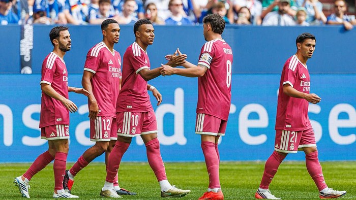 Five soccer players in pink uniforms celebrate on a green field, exchanging handshakes as fans watch from the blue advertising boards in the background.