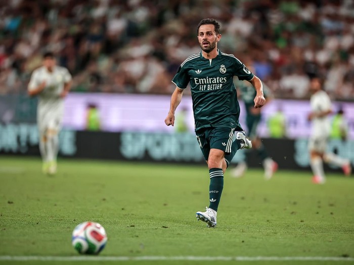 A soccer player in a dark green Real Madrid kit runs toward a ball on a grass field during a match, with a blurred crowd in the background.