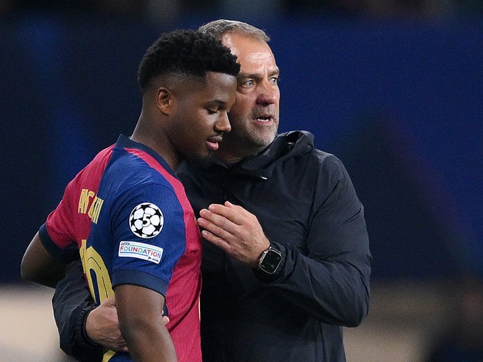 Soccer coach in a dark jacket hugs a young player in a red-and-blue jersey on the field after a match, signaling support or consolation.
