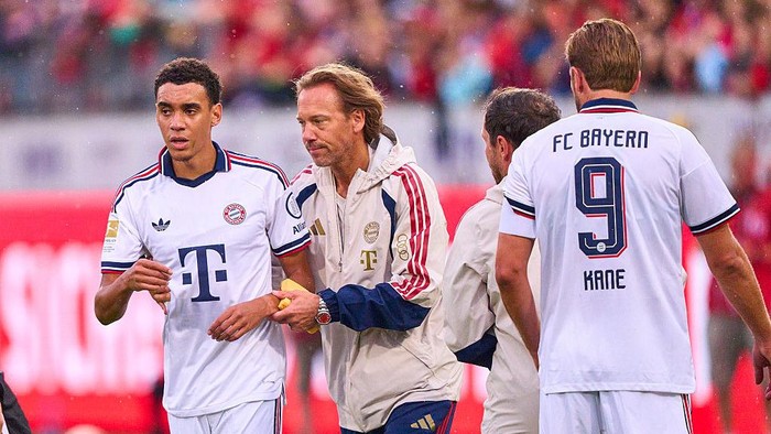Three soccer players in white Bayern Munich kits on the field; one has '9 KANE' on his back and a coach walks with them.