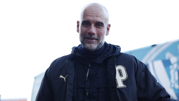 A bald man with a gray beard wearing a dark jacket with a large letter P on the chest, standing outdoors with a blue structure in the background.