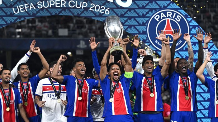 Paris Saint-Germain players in blue and red jerseys celebrate on stage, lifting the UEFA Super Cup trophy as confetti falls, with the PSG crest visible in the background.