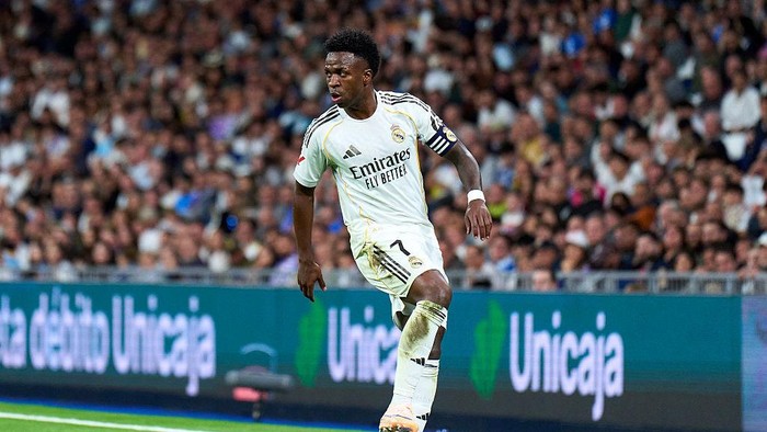 Soccer player in a Real Madrid white kit dribbling the ball with a crowded stadium behind him, mid-action on the pitch.