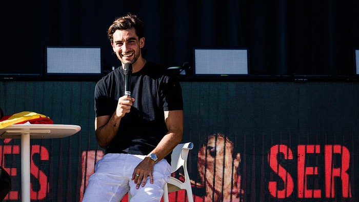 Smiling man in a black t‑shirt and white pants sits on a stool on stage, holding a microphone during a talk, with a large LED screen behind him.