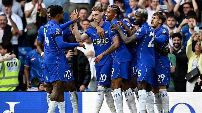 Soccer players in blue uniforms celebrate a goal on the field as fans cheer in the stands behind them.
