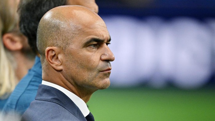 Side profile of a bald man in a suit with a serious expression on a sports field backdrop