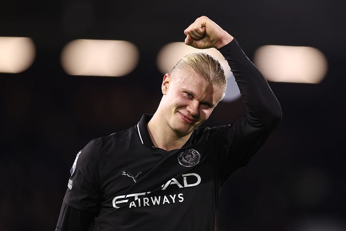 Soccer player in a black kit raising a clenched fist in celebration on the pitch, smiling at the camera with a dark background.