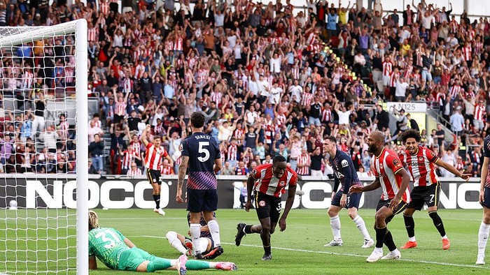 Soccer match near the goal: goalkeeper on the ground, ball loose, players in red-and-white stripes and dark kits nearby as the crowd cheers in the stands.