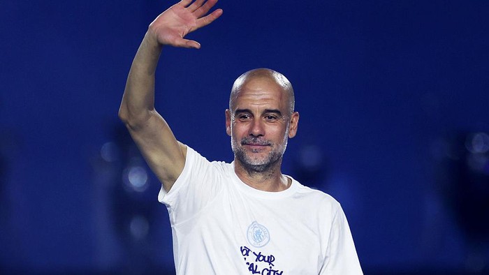 Man with a shaved head in a white T‑shirt waving to the crowd on a blue-lit stage.