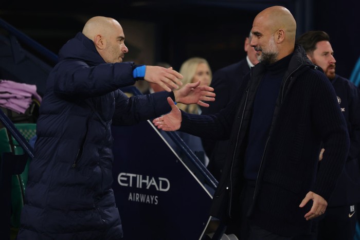 Two bald men shake hands on a sports sideline, one in a navy puffer jacket and the other in a dark sweater, with Etihad Airways signage in the background.