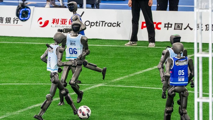 Humanoid robots playing soccer on a green field with numbered bibs 06 and 05, competing near banners in the background.