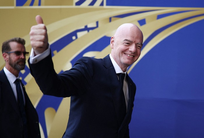 Smiling bald man in a dark suit gives a thumbs up as he walks past a blue and yellow backdrop with abstract shapes.