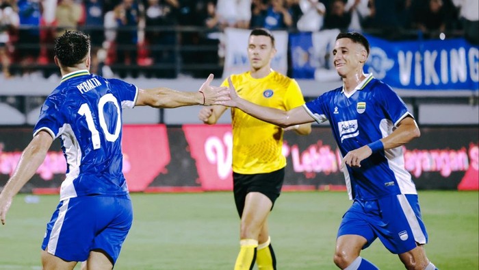 Two soccer players in blue jerseys celebrate with a high-five on the field as a yellow-clad referee watches.