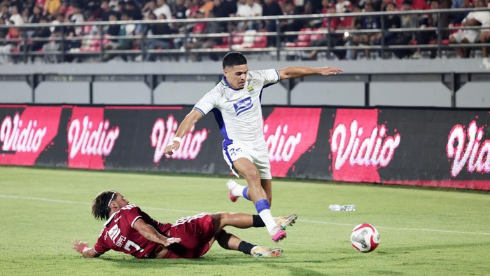 Soccer match: white-jersey player leaps over a sliding tackle as a ball rolls nearby on the field, with spectators watching from the stands.