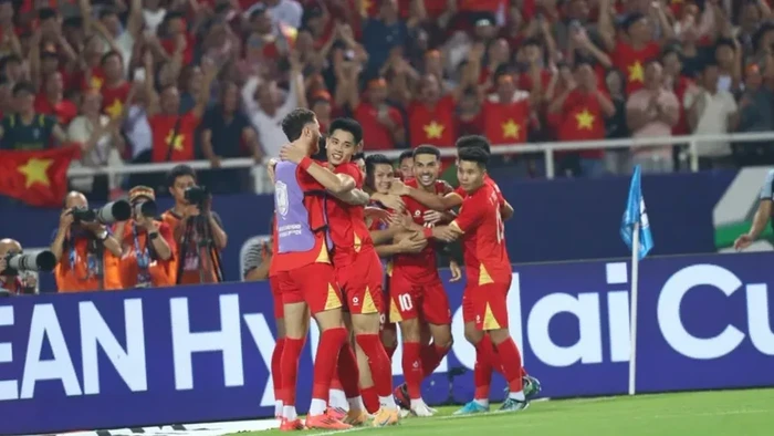 Group of red-uniform soccer players celebrate together on the field as fans with Vietnamese flags cheer in the stands.