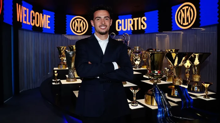 Man in a dark suit stands with arms crossed in a trophy-filled backstage area, with a 'Welcome Curtis' sign in the background.