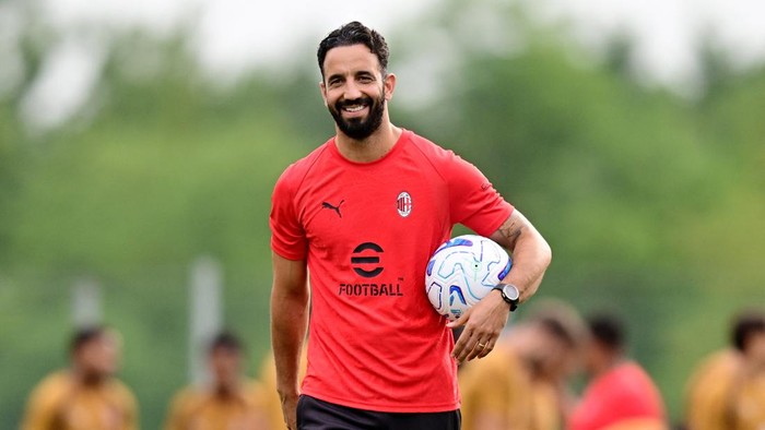 Athletic man in a red Milan training shirt holding a white soccer ball on a practice field, smiling on camera.