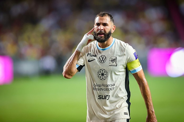 Soccer player wearing a white jersey with a yellow captain's armband, gesturing by his ear on the pitch during a match.