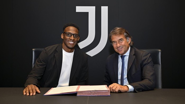 Two men sit at a dark conference table signing a document, with a large Juventus logo on the wall behind them.
