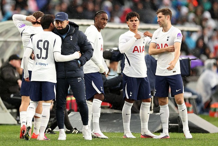 Tottenham Hotspur players in white jerseys stand on the sideline, listening to a coach during a rainy match context.
