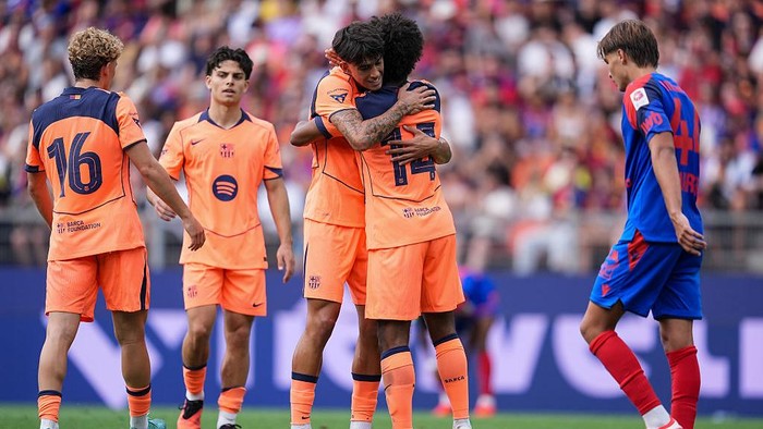 Two players in bright orange jerseys hug on a soccer field as teammates stand nearby and a blue-clad opponent looks on.