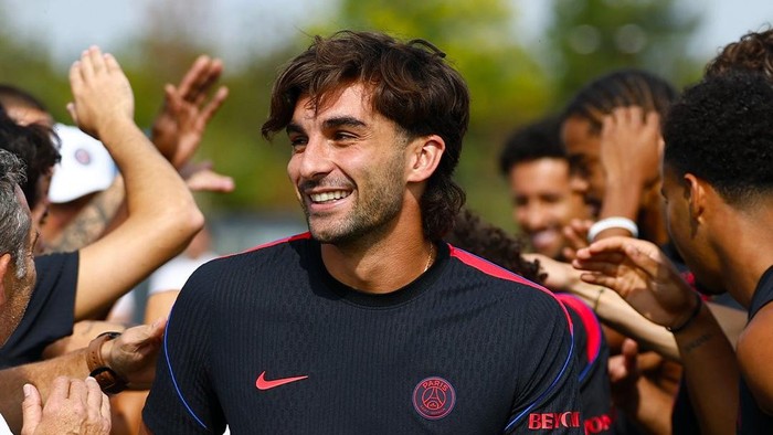 Soccer player in a PSG training jersey smiling as teammates celebrate with high-fives around him.