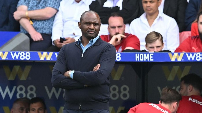 Football manager standing with arms crossed in the dugout during a game, spectators watching from the stands.