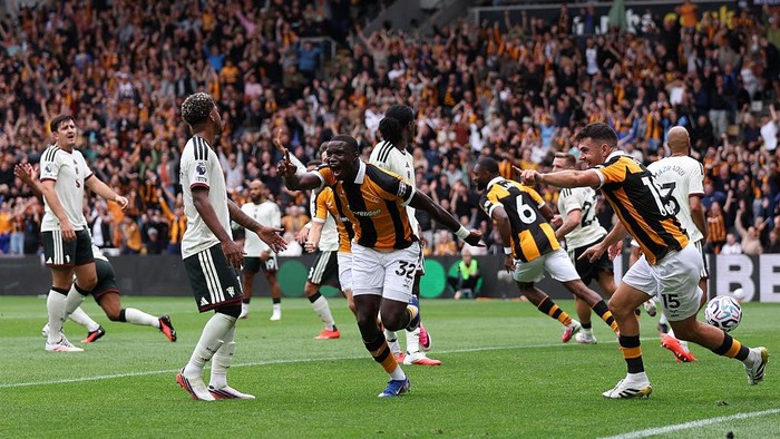 Soccer players in orange and black celebrate a goal on a crowded stadium field, with a ball near the right and fans cheering in the stands.